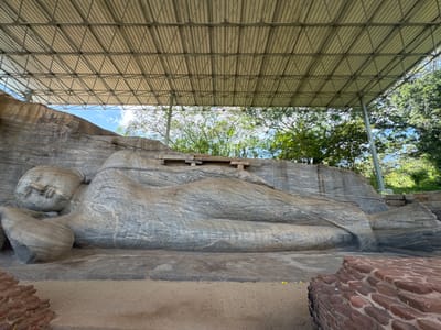 The image shows the reclining Buddha statue at Gal Vihara in Polonnaruwa, Sri Lanka. The statue is carved from a large granite rock face and depicts Buddha lying on his right side. The statue is under a modern protective shelter with a metal framework and roof. In the background, there are trees and a glimpse of the sky. In the foreground, there is a sandy area and a section of brickwork.