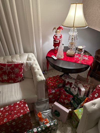 A room displays a decorated scene with Christmas gifts. A white tufted sofa is adorned with a red snowflake pillow. A round table, draped with a red runner, features glass decorations and a harlequin doll. Numerous wrapped presents with holiday-themed paper are scattered on the floor near the sofa, adding to the festive atmosphere. A table lamp illuminates the setting, complementing the ambiance.