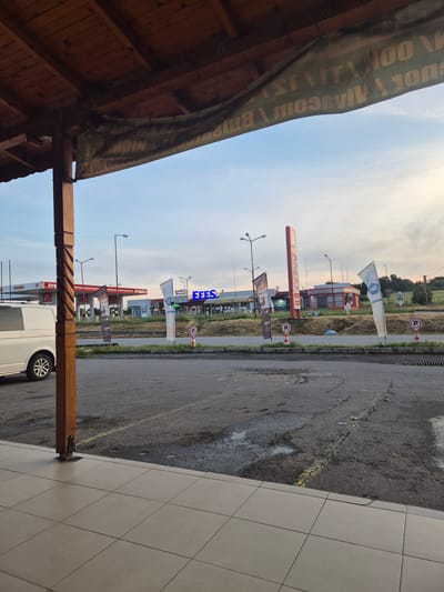 Here's a description of the image:
The image shows a view of a gas station in Lesovo, Bulgaria, from under a wooden structure. The gas station has the logos of Lukoil and FEES visible. Several flags and signs are present along the side of the road. A white van is partially visible in the foreground. The ground is a mix of pavement and tile. The sky is visible in the background.