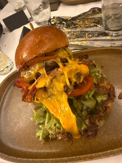 A hamburger, impaled with a toothpick, sits on a brown, textured plate beside mixed greens. The burger is topped with melted cheese, tomato slices, and bacon. The plate is placed on a table with napkins and glassware. The burger bun appears slightly toasted.