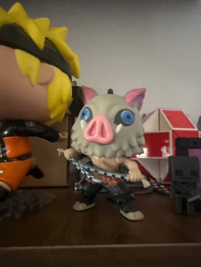 This is a close-up, eye-level shot of several collectible figurines displayed on a dark brown wooden surface. The foreground is dominated by a character wearing a pig mask with blue eyes and a pink snout, holding a chain weapon. To the left of this figure is another figurine, partially visible, with bright yellow hair and an orange and black outfit. In the background, behind the pig-masked figure, are two more figurines: one is a dark grey, blocky character with a purple accent, and another is a red and white geometric structure. The lighting suggests an indoor environment, possibly a shelf or display case, with a light grey wall in the background. The image does not provide any information about the city of Leominster, United Kingdom.
