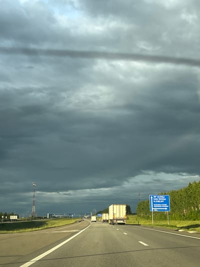 The image shows a highway in the vicinity of Kreshchenye Kazyli, Russia. The sky is heavily overcast with dark, dramatic clouds, suggesting an impending storm. A tall communication tower is visible on the left side of the road, and a line of trees borders the right side. Several trucks are traveling on the highway, with one white semi-trailer truck prominently featured in the foreground. A blue road sign with white lettering provides directions to "ЯР ЧАЛЛЫ," "НАБ.ЧЕЛНЫ," and "N.CHELNY," along with other locations and a distance marker of "300м." The road itself is asphalt with white lane markings. The scene is captured from the perspective of a vehicle traveling on the highway.