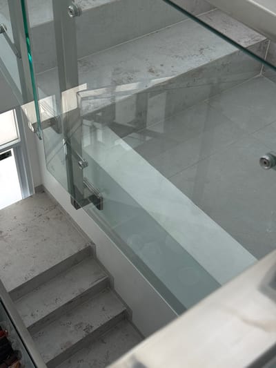 The image displays a modern interior staircase with glass railings. The steps and landings are tiled with a grey, textured material. A vertical glass panel forms the railing, supported by metal brackets. The lighting suggests an indoor environment. The image does not provide any contextual clues to suggest the location is in Tanjungmeru, Indonesia.