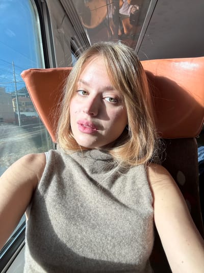 The image is a close-up selfie of a young adult female taken inside a train car. She has blonde, shoulder-length hair with bangs, green eyes, and wears light pink lipstick on her full lips. Her attire consists of a beige sleeveless turtleneck top. Direct sunlight from the left illuminates her face and casts shadows. Behind her, an orange upholstered train seat is partially visible. Through the train window to her left, a clear blue sky, railway electrical wires and poles, and distant buildings with terracotta roofs on a green hillside are discernible. This external view is consistent with a railway line within the city of Ventimiglia, Italy.