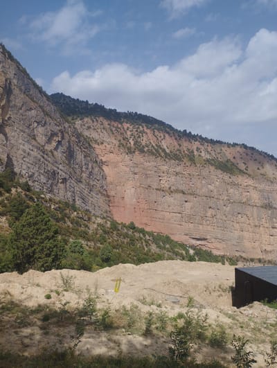 The image depicts a vast, layered rock face of a mountain under a cloudy blue sky. The mountain exhibits distinct geological strata, with varying colors ranging from gray to reddish-brown, indicating different rock compositions. Sparse vegetation, including trees and shrubs, grows along the lower slopes and at the base of the mountain. In the foreground, a sandy area with some scattered plants and shadows suggests a somewhat disturbed or undeveloped landscape. A dark, modern-looking structure with a slanted roof is partially visible on the right side of the frame. The overall scene is natural and rugged, with a single man-made structure present. The location is identified as Chaknak, Uzbekistan.