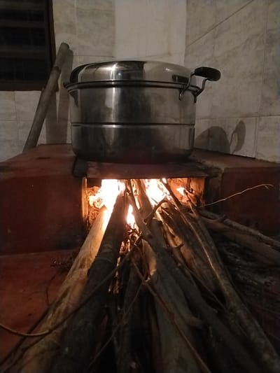 A rustic outdoor cooking scene unfolds in Laranjal Paulista, Brazil, where a large, multi-tiered stainless steel steamer pot rests atop a brick stove. The pot is positioned directly over a vibrant, crackling fire, fueled by a substantial pile of dry, elongated logs arranged in a pyramid formation. The fire's intense heat emanates from within the stove, illuminating the surrounding environment with a warm, orange glow. To the left of the stove, a wooden pole leans against the tiled wall, its purpose unclear. The background features a tiled wall, suggesting an outdoor kitchen or a simple cooking area. The image captures a moment of traditional cooking, highlighting the use of fire and natural materials in food preparation.
