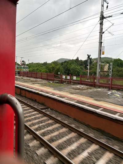 A train moves past a station on a cloudy day. The perspective is from within the train, looking out a window. A portion of a red train car is visible on the left, obscuring a portion of the view. In the distance, a green and white train engine is stopped on the tracks. A red fence lines the platform of the station. Overhead, power lines crisscross the sky. Lush green trees and hills form the background.
