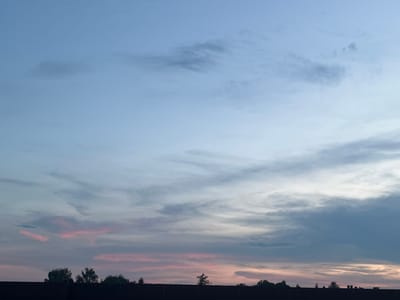 The image depicts a dusky sky with varying cloud formations over the city of Ożarów Mazowiecki, Poland. The upper portion of the sky is a pale blue, transitioning to lighter shades near the horizon. Wisps of grey and darker grey clouds are scattered across the sky, with some appearing more layered and textured. Near the horizon, the clouds are tinged with soft pink and peach hues, suggesting the last light of sunset or the approach of dawn. Silhouetted against this sky is the dark, horizontal line of rooftops and trees that form the city's skyline. The trees appear as dark green shapes with indistinct foliage. The overall impression is a serene, natural scene observed from an elevated vantage point within the urban landscape of Ożarów Mazowiecki.
