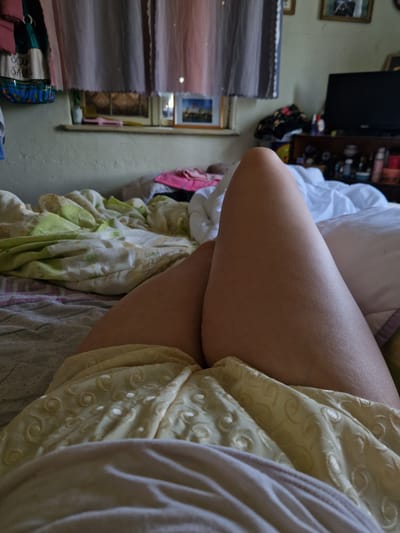 The image depicts a close-up view of a person's legs, resting on a bed. The person is lying on their back, with their legs bent at the knees and crossed at the thighs. They are wearing a yellow garment with a subtle pattern of swirls and circular shapes. The bed is covered with various blankets and sheets, some with floral patterns in green and white, and others in muted grey and white. In the background, a window is visible, adorned with sheer curtains in shades of pink and gray. The windowsill holds a few small objects, and beyond the window, a glimpse of an outdoor scene is suggested. To the right of the window, a television set is visible on a dark wooden shelf unit, along with various items on the shelves. The overall environment appears to be a bedroom, possibly in a residential setting. The location is not specified in the image.