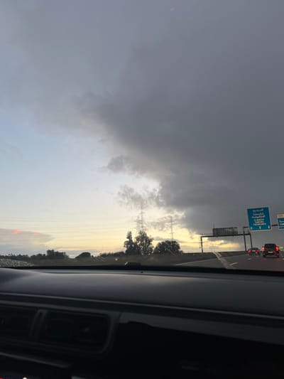 The photograph shows a cloudy sky from inside a vehicle. Dark storm clouds fill the left side of the frame. Power lines and a metal transmission tower appear in the distance on the right, with trees below. A road sign is visible above the car in front. The overall light suggests it may be either sunrise or sunset.