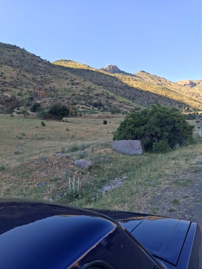 This media file shows a rural, mountainous landscape with a clear blue sky. The image is oriented vertically, with the sky occupying the left half and the terrain filling the right. A dark blue car is parked on the right side, its polished surface reflecting the mountainous scenery. The mountains themselves are rugged and covered in dry grass and sparse vegetation, with some rocky outcrops visible. A large, solitary tree stands prominently in the middle ground, next to a large boulder. The sun appears to be low in the sky, casting long shadows and highlighting the texture of the mountain slopes, suggesting it is either early morning or late afternoon. The overall scene conveys a sense of peaceful solitude in a natural, undeveloped area. No people or text are visible. Given the location context of Shatin, Armenia, the scene depicts a remote and arid mountain environment characteristic of the region.