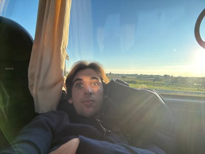A young male subject with light brown hair and a surprised expression, characterized by wide-open eyes and a slightly agape mouth, is depicted in a selfie-style photograph. He is wearing a dark blue long-sleeved garment and appears to be seated inside a vehicle, likely a bus or coach. To the subject's left, a dark-colored seat headrest is visible, bearing the white text "MAN." Adjacent to the seat is a beige fabric curtain or blind. The right side of the image is dominated by a large window overlooking a bright, clear blue sky. The sun is low on the horizon to the right, creating significant lens flare and glare on the window glass. Beyond the window, the landscape consists of expansive, flat green fields, with faint silhouettes of structures or trees visible in the distant horizon line. The scene is illuminated by natural sunlight, suggesting either early morning or late afternoon. The location depicted is within or in transit through the city of Ladispoli, Italy.