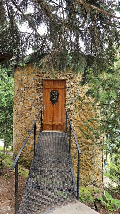 A weathered wooden door is set into a round tower constructed of rough-hewn, honey-colored stones. The door is adorned with a black, shield-shaped emblem featuring a circular design and the Cyrillic word "БОТАШ" beneath it. A metal ramp with black railings leads up to the entrance. The structure is surrounded by lush green trees and foliage, with a dense canopy of pine branches overhead partially obscuring the sky. The scene suggests a rustic, possibly historical or themed, building within a natural setting, likely in the city of Krasnyy Kurgan, Russia.