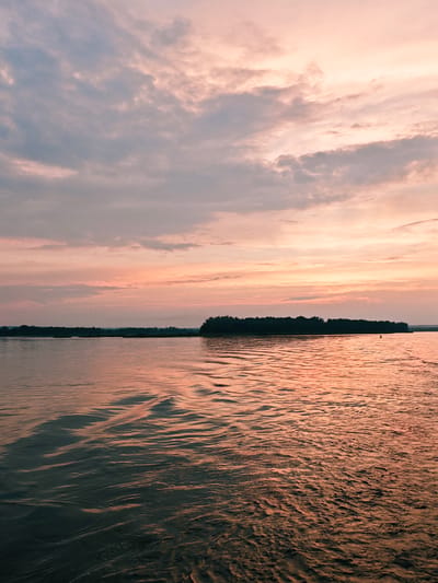 The image depicts a serene sunset over a wide body of water, likely a river or lake, near Nakhval'skoe, Russia. The sky is a canvas of soft pastel colors, transitioning from pale blue at the top to hues of pink and orange closer to the horizon, with scattered clouds. A dark, dense line of trees forms a natural border across the distant shore. The water's surface is gently rippled, reflecting the warm tones of the sky and appearing darker in the foreground. The scene is peaceful and devoid of any human presence or discernible activity.