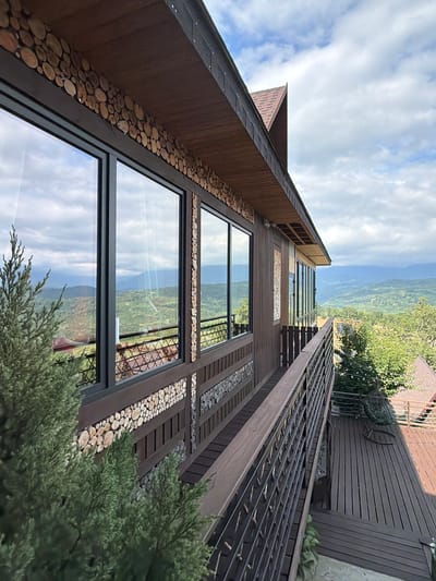 This is a slightly high-angle, eye-level outdoor shot of a modern wooden building with large windows, situated on a hillside with a view of a valley and distant mountains. The building's exterior is decorated with rows of circular wood slices. A dark wooden balcony with a metal railing runs along the side of the building, overlooking a wooden deck. In the foreground, a portion of a green evergreen tree is visible. The sky is partly cloudy. The scene appears to be located in Nizhnyaya Shilovka, Russia, given the architectural style and natural landscape. There are no people or animals visible in the image.