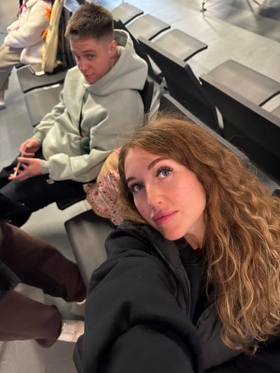 The image appears to be a selfie taken inside an airport waiting area. A young woman with long, curly blonde hair and blue eyes is facing the camera, wearing a black jacket. To her left is a young man wearing a light green hoodie. Both are seated on black airport-style seating. In the background are rows of empty chairs. The location is not Boldino, Russia.