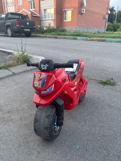 A red toy motorcycle sits on an asphalt road in front of a brick apartment building in Gukovo, Russia. The motorcycle is facing the camera and its front wheel is large and worn. The building in the background has several windows, some of which reflect sunlight. A black pickup truck is parked on the left side of the image, near the building. The area around the building appears to be a residential street with some greenery.