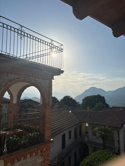The image depicts a sun-drenched view from a balcony in Orino, Italy, overlooking a valley dotted with tiled-roof houses and distant mountains. The foreground features a decorative wrought-iron balcony railing and a red brick archway. The sun's glare is strong, creating a bright halo effect in the sky. The midground shows several residential buildings with terracotta roofs, and the background consists of layered mountain ranges fading into a hazy blue sky. Lush green trees are visible, adding to the scenic landscape. The overall scene is tranquil, showcasing a typical Italian hillside village bathed in the warm light of day.