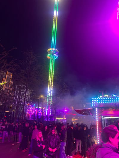 The image depicts a nighttime scene at a fair in Groningen, Netherlands. A tall amusement ride with a central column of light and spinning, ring-shaped attachments at the top is illuminated with colorful green, yellow, and blue lights. A crowd of people stands in the foreground observing the ride. There are also trees. Further back, another amusement ride is visible, featuring a vertical display of lights. The overall atmosphere is lively and festive.