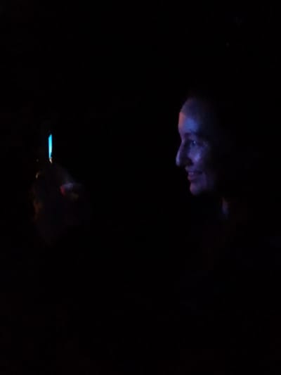 In a dark environment, a person's profile is visible on the right side of the image, illuminated by blue and purple light. Their face is turned to the left, with their mouth slightly open in a smile. On the left side, a hand holds a small object that emits a bright blue light. The background is completely black, suggesting the scene is set at night or in a dimly lit indoor space. The image does not provide any specific location in Schwerin, Germany, or any details that would suggest it.