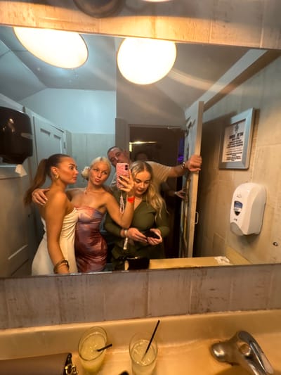 A mirror reflects five people posing for a photo in a bathroom. The reflection shows three women in the foreground, one in a white dress, another in a purple-toned dress, and the third in a green top, holding a phone. Two other people, a man and a woman, are further back. The bathroom features a soap dispenser, a framed sign, a sink with a faucet, and two cocktails with straws on the counter. The room is illuminated by bright overhead lighting.