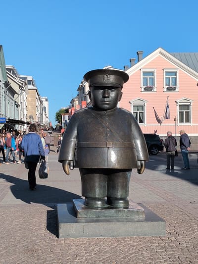 In Oulu, Finland, a large bronze statue of a stout man in a uniform with a hat stands prominently in the foreground of a pedestrian street. The statue faces forward and is positioned on a grey stone base. Behind the statue, a pale pink building with white trim and symmetrical windows is visible, adorned with flags. The street is paved with cobblestones, and several people are walking in the background. To the left of the statue, a woman in a blue jacket carries a shopping bag. A dark SUV is parked on the right side of the street. The sky above is clear and blue.