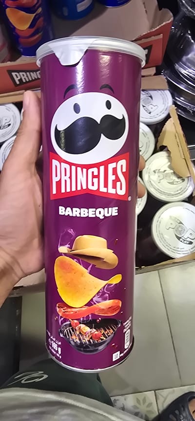 A person's hand holds a Pringles BBQ flavor potato chip can. The can is purple with the Pringles logo and an illustration of potato chips, hot dogs, and a grill. The background is a bit blurry, showing other Pringles cans, some plastic containers, and what appears to be a box. The setting seems to be indoors, possibly a pantry or storage area. The lighting is bright, suggesting daytime. There is text visible on the can: "PRINGLES" and "BARBEQUE." The overall impression is casual, possibly someone is showing off a snack they have.