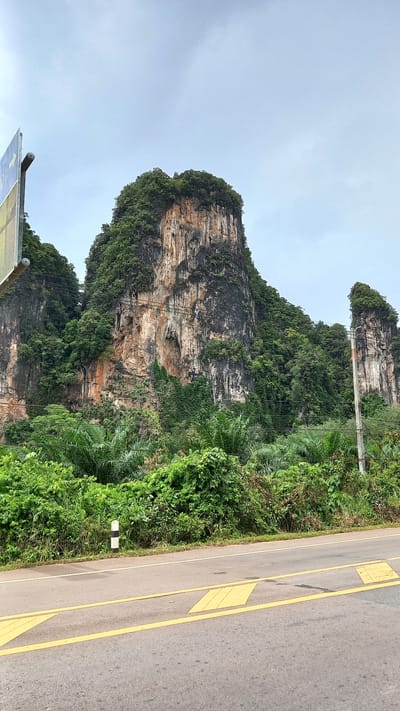 The image depicts a large, rocky cliff face covered in lush green vegetation in Khao Khram, Thailand. The cliff has a layered appearance with exposed rock faces of orange and gray. The base of the cliff is obscured by dense foliage. A road runs along the right edge of the frame, marked with yellow lines and painted speed bumps. The sky is overcast and a pale gray color. Power lines are seen running from the top of one of the cliff sides. A sign is visible to the left in the lower portion of the frame.