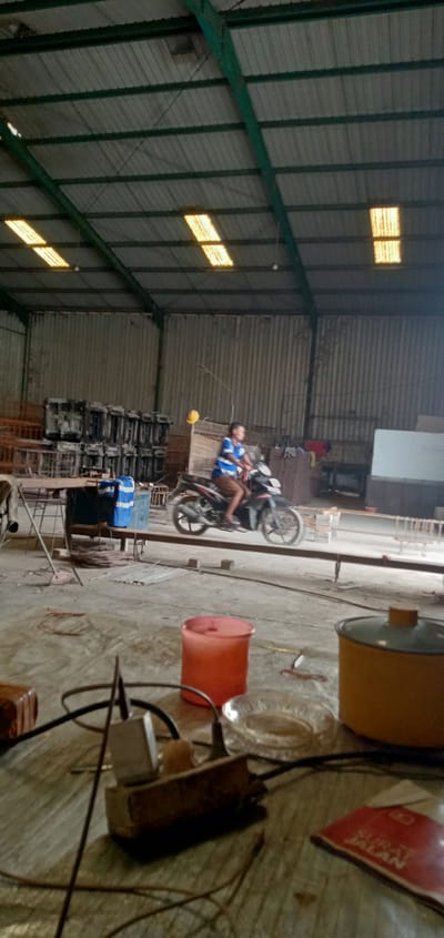 The photo appears to be taken inside a warehouse or industrial building in Sumput, Indonesia. A man is riding a black and silver motorcycle on a platform within the building. The ceiling is high and constructed of metal, with intermittent translucent panels allowing light to filter through. To the left, there are stacks of gray, rectangular objects, possibly concrete forms, and other metal structures. The foreground shows a cluttered table with electrical cords, an orange plastic container, a yellow pot, an ashtray, and a red package labeled "SURAT JALAN." The building has corrugated metal walls.