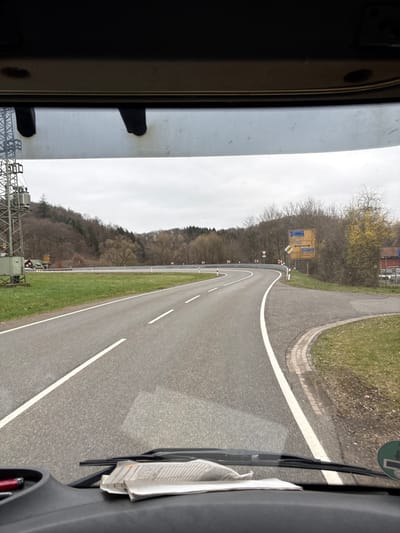 The image displays a forward view from inside a vehicle on Bundesstraße 41 (B41) approaching the junction with Landesstraße 135 (L135) in the municipality of Nohfelden, Germany, specifically near Türkismühle. The asphalt road curves right in the foreground with solid white edge lines and broken white center lines. To the right, a smaller asphalt road branches off, edged with a paved, brick-bordered shoulder. A large yellow directional sign indicates "Eisen" and "Sötern" straight ahead, and "Nohfelden" and "Türkismühle" to the right. Multiple red-bordered white curve warning signs are positioned along the right side of the main road, indicating a right turn. Further right, behind the signs, a building with a red roof and a visible parking area is present. On the left side of the road, a metal lattice structure, possibly a power transmission tower, stands next to the road. A small building with a red roof is visible behind this structure. The surrounding environment consists of rolling hills covered with bare deciduous trees under an overcast sky, indicating late autumn or early spring. Inside the vehicle, a crumpled piece of paper, possibly a receipt or note, rests on the dark dashboard. A wiper blade is partially visible, and a circular green sticker is affixed to the lower right of the windshield. No people are visible.