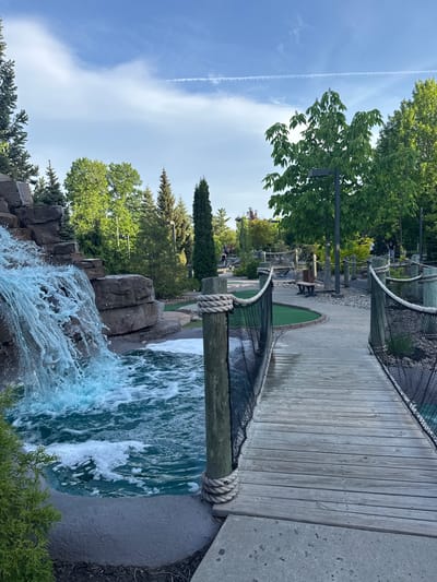 The image depicts a vibrant outdoor recreational scene, likely a mini-golf course, under a clear, bright blue sky with a wispy white contrail. In the foreground, a sturdy wooden bridge with rope and black netting railings spans over a man-made waterfall. The waterfall features foamy, blue-green water cascading over textured artificial rocks.

Beyond the bridge, the mini-golf course is visible, featuring a green turf hole set into a sandy path. The course extends into the background with various elements such as benches, additional rock formations, and more lush greenery. Several indistinct figures can be seen in the distance, suggesting people are engaged in playing mini-golf.

The area is richly forested with a mix of tall coniferous trees and leafy deciduous trees, providing ample shade and a natural aesthetic. A dark metal light post stands among the trees. The time of day is bright and sunny, indicating midday or early afternoon, contributing to the pleasant outdoor atmosphere. The overall setting suggests a well-maintained leisure facility designed for outdoor recreation in Chelsea, Canada. No visible text is present in the image.