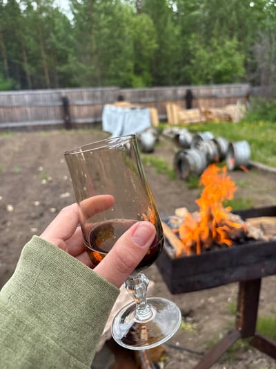 A person holds a glass of dark liquid, possibly wine or beer, with their left hand. They are wearing a light green long-sleeved shirt. In the background, a fire burns in a black barbecue grill. Beyond the grill, there is a wooden fence, some scattered lumber, and what appear to be stacked metal barrels. The scene is outdoors, with trees and greenery visible in the distance. The ground appears to be dirt and gravel. The location is not specified within the city of Zheleznodorozhnyi, Russia.