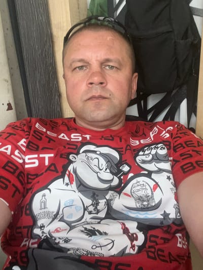 A vertical, slightly angled medium shot captures a man from the chest up, positioned in a dimly lit, indoor setting. He has light skin, brown eyes, and short, dark hair. He is wearing a red t-shirt with a repeating pattern of black and white text and graphic designs, including what appear to be cartoonish fists with tattoos and images of a sailing ship and dice. The man is looking directly at the camera, his expression neutral. In the background, a dark, possibly black, fabric item is visible to his left, and a striped surface, perhaps a piece of furniture, is to his right. The lighting is dim, suggesting it might be evening or an indoor environment with low light. There is no clear indication of weather or a specific time of day beyond that. The text on the shirt is partially visible and appears to be in English, with some words like "BEAST" and "EAST" discernable.