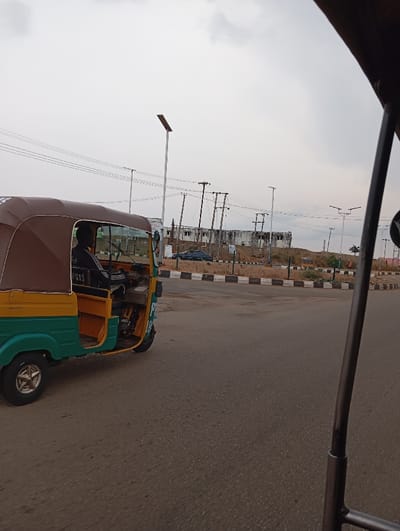 The image, taken in Dadin Kowa, Nigeria (latitude: 9.813, longitude: 8.866), shows a green and yellow auto-rickshaw, known locally as a “Keke,” driving on a paved road. The Keke is moving away from the viewer and has a brown canopy top. A person is driving the Keke. The driver is wearing a black shirt. There is a power pole and a streetlight to the left of the Keke, and a white building with a brown roof in the background. There is a black car parked on the left side of the road, near the building. The Keke is driving on a slight curve to the right. The road has white lines and black painted curbs. The weather is overcast. The ground is dry and dusty.