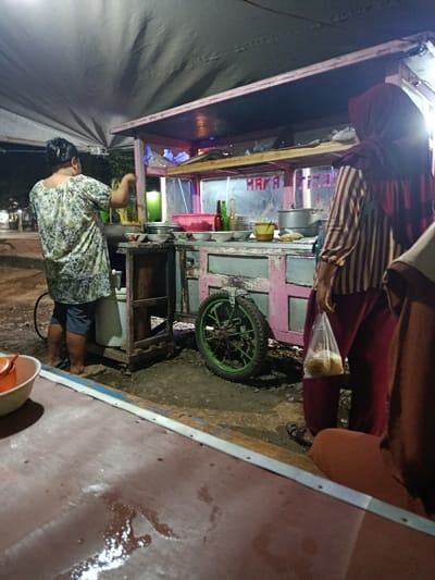 This image depicts a nighttime street food vending operation in Arjasa, Indonesia. The scene is illuminated by artificial light sources under a grey canvas tarp that serves as a canopy.

In the center, a pink, wooden, wheeled food cart with glass display sections is situated on unpaved ground. The cart features a prominent green-spoked wheel on its right side. On its shelves, various items are arranged, including white bowls, clear containers holding reddish ingredients, several bottles, and a yellow mug. Red lettering spelling "MAYA TIZAN" is visible on the cart's front glass panel.

On the left, a person, viewed from behind, is engaged in preparing food. This individual wears a light-colored, patterned tunic and dark shorts, with hair pulled back. They are positioned over a cooking vessel on the cart, holding a utensil in their right hand. A white container is visible on the ground next to their feet.

On the right, another person, also viewed from behind, stands near the cart. This individual wears a striped long-sleeved shirt, dark-red pants, and a dark-red headscarf. They are holding a clear plastic bag containing light-colored items, likely food, in their left hand.

In the foreground, a reddish-brown flat surface, possibly a table or counter, occupies the lower portion of the frame. A white bowl with an orange-spouted container rests on this surface in the lower-left, and liquid spills are visible on its surface. The ground beneath the food cart is irregular, appearing to be dirt or gravel. The background is dark and blurred, with distant lights suggesting an urban environment at night.