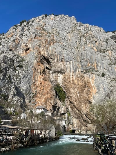 The image depicts a dramatic natural and built landscape at Blagaj, Dračevice, Bosnia and Herzegovina, centered on the Blagaj Tekke complex. A colossal, rugged limestone cliff, exhibiting shades of grey, white, and prominent rust-orange/brown geological streaking, dominates the background, rising steeply to meet a clear blue sky. Numerous fissures, cracks, and small caves are visible on the cliff face, with sparse green vegetation clinging to its upper ledges and crevices.
At the base of the cliff, a powerful karstic spring emerges from a large, dark cave opening, forming turbulent white rapids as it flows into the turquoise-green Buna River, which occupies the foreground. On the left bank, a cluster of traditional Ottoman-style stone buildings with white facades and grey slate roofs is nestled directly against the cliff face. The most prominent structure, likely the Tekke, features multiple windows and a distinct roofline.
Bare deciduous trees with intricate branch structures line both riverbanks. On the left, an outdoor dining patio with tables and chairs under a canopy extends over or adjacent to the river. A similar outdoor seating area, partially obscured by signage, is visible on the right bank. No human figures, vehicles, or active interactions are visible in the scene, which is illuminated by natural daylight.