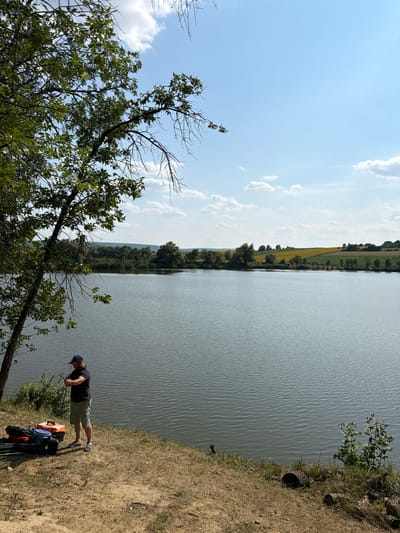 A man is fishing on the bank of a lake. He is wearing a dark t-shirt, light-colored shorts, a baseball cap, and sandals. He is holding a fishing rod. Several fishing tackle boxes and equipment are on the ground next to him. The lake is calm with a few ripples on the surface. The background shows a line of trees and rolling hills with fields of yellow flowers, possibly sunflowers. The sky is blue with scattered white clouds. The scene is set in Pănăseşti, Moldova.