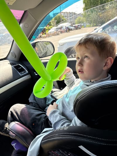 A young child is seated in a car seat, inside a vehicle, holding a green balloon animal in the shape of a flower or clover. The child is wearing a grey sweatshirt and dark pants. The vehicle appears to be parked, with a view of a street and buildings visible through the window. There's no evidence to suggest this image was taken in the city of Италмас, Russia.
