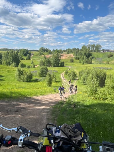 This outdoor daytime image, captured in the vicinity of Novochnuvashsky, Russia, depicts a group of cyclists navigating a series of unpaved dirt paths through a verdant, rolling landscape under a bright blue sky with scattered white cumulus clouds.

In the distant background, gentle hills are covered with dense deciduous forests, with a few white residential-style buildings topped with dark roofs visible on a distant crest. Far left, faint industrial structures or power lines are discernible.

The midground is characterized by expansive green fields interspersed with patches of purple and yellow wildflowers. Numerous small, bushy deciduous trees are scattered throughout the fields and along the winding dirt paths. A group of approximately ten individuals, primarily on mountain bikes, are seen traversing these paths, generally moving away from the viewer. One cyclist in the foreground group wears a blue jacket and stands next to a yellow mountain bike. Other cyclists are visible further along the paths, navigating turns and inclines.

The image is taken from the first-person perspective of a person on a motorcycle or dirt bike, with the vehicle's handlebars prominently featured across the bottom of the frame. Visible components include a silver-colored clutch lever assembly on the left, black rubber grips, and left-hand controls with red, blue, and gray buttons/switches. A pair of off-white and blue patterned gloves or a cloth, bearing a "100%" logo, is draped over the right side of the handlebars. The immediate foreground is a dirt track, leading into the broader path network.