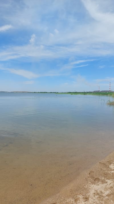 A tranquil scene unfolds at the edge of a large body of water, likely the Topar Reservoir, near the city of Topar, Kazakhstan. The foreground features a sandy beach, lightly textured with scattered pebbles and a few footprints pressed into the damp sand, indicating recent human presence. Beyond the beach, the calm, clear water stretches out, revealing the sandy bottom in shallower areas. The middle ground is characterized by the vast expanse of water, reflecting the sky and distant elements. Along the far bank, a dense line of reeds and other vegetation creates a natural border. In the distance, industrial structures are visible: two prominent red-and-white striped chimneys rise from a large building, emitting a faint plume of smoke, suggesting a power plant. Other utility poles and structures are scattered along the horizon. The sky above is a bright blue, adorned with wispy white clouds that add a sense of depth and movement to the scene. The overall atmosphere is one of calm, with a juxtaposition of natural landscape and industrial activity.