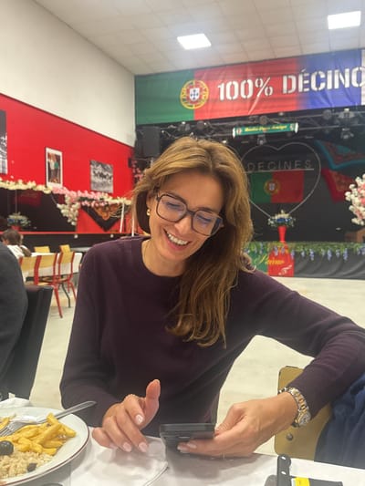 The image captures a smiling woman seated at a table in an indoor venue located in Décines-Charpieu, France. The woman has medium brown hair, wears tortoise-shell framed glasses, and is dressed in a dark purple long-sleeved top. She is actively looking down at a smartphone, encased in black, which she holds with both hands. A silver-toned watch with a metallic band is visible on her right wrist, and her fingernails are painted light pink. A white plate positioned in the foreground to her left contains french fries, a grain-based dish (possibly rice or couscous), and several black olives, alongside a fork and knife.
The background reveals a large event space with a stage area. Dominant decorations include large banners: one featuring the Portuguese flag and the text "100% DÉCINES", and another with "DÉCINES" inscribed within a heart shape, also incorporating the Portuguese flag. Further banners display "Maísa Doce Amigo" and "J'aime" with floral accents. The walls are painted red on the lower section and white above, adorned with garlands of pink and white artificial flowers. A tall red vase holding blue flowers stands on the stage. Square ceiling lights illuminate the space. In the distance, multiple empty tables with yellow and red chairs indicate a dining or event setup. A dark-clothed arm of another individual is partially visible in the left foreground.