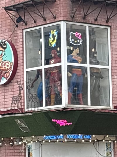 The image depicts a storefront in the city of Tutaev, Russia. The building features a brick facade and a large corner window display. Inside the window, there are life-sized figures of comic book characters, including Superman, Flash, and Batman. Neon signs in the shape of Hello Kitty and a yellow lightning bolt are also visible in the window. Outside the window, there are decorative metal reindeer figures. Below the window, a green awning is adorned with a logo and string lights. Further down, another neon sign reads "ГОРОДСКОЕ КАФЕ" which translates to "CITY CAFE". The overall scene suggests a themed cafe or shop.