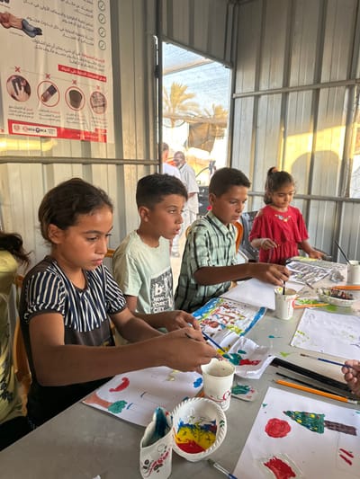 The image depicts an indoor art activity within a temporary or modular structure in a-Zawayda, Palestinian Territory. Four children, estimated to be between 6 and 12 years old, are seated at a light-colored table, engaged in painting.

From left to right:
1.  A girl, approximately 10-12 years old, with dark hair and wearing a dark shirt with vertical white stripes, holds a paintbrush and focuses on her paper.
2.  A boy, approximately 9-11 years old, with short dark hair and wearing a light blue t-shirt with a "NEW YORK CITY" graphic, looks towards the right while holding a paintbrush.
3.  A boy, approximately 9-11 years old, with short dark hair and wearing a green and white plaid shirt, holds a paintbrush and concentrates on his artwork.
4.  A girl, approximately 6-8 years old, with dark hair tied up and wearing a red off-the-shoulder top, is focused on her painting with a paintbrush.

The table is covered with white sheets of paper, some displaying colorful drawings including trees and abstract shapes. Various art supplies are distributed across the table, including plastic cups holding water or paint, small white bowls containing mixed paints (red, yellow, blue), and multiple paintbrushes.

The interior walls and ceiling of the structure are constructed from corrugated metal. A white banner with Arabic text, safety graphics, and "DCA" and "NCA" logos is affixed to the metal wall on the left. A large opening on the right side of the structure allows natural light to enter and offers a view of the outdoors. Visible outside are several palm trees and what appear to be tents or temporary structures. Two adult males are partially visible in the background, standing outside the structure; one wears a white shirt, the other a light-colored traditional garment. The children are individually focused on their painting activities, exhibiting no direct interaction with each other or the background figures.