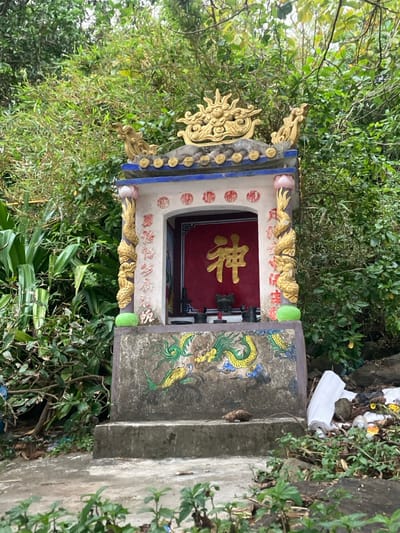 An outdoor scene in Sơn Trà Ward, Vietnam, depicts a small, ornate shrine situated amidst dense green foliage. The shrine features a multi-tiered concrete base with two steps. A painted green and yellow dragon motif with blue cloud accents adorns the front of the base. A small, brown rodent is present on the second step of the base, positioned towards the right. The shrine's main structure is white with gilded decorative columns featuring dragon figures and lotus motifs at the corners. The central recess contains a red background with a prominent gold Chinese character "神" (Shén, meaning 'deity' or 'spirit'). Within this recess, two dark, metallic-appearing vessels or incense burners are visible. The shrine is topped with a blue and white tiled roof, featuring a large, golden, ornate finial resembling a dragon head. Dense green vegetation, including broad-leaf plants and bamboo, forms the background and flanks the shrine. To the right of the shrine, a accumulation of assorted waste, including white plastic and bottles, is visible on the ground. The lighting is diffused, suggesting an overcast day or shaded location. No human subjects are present.