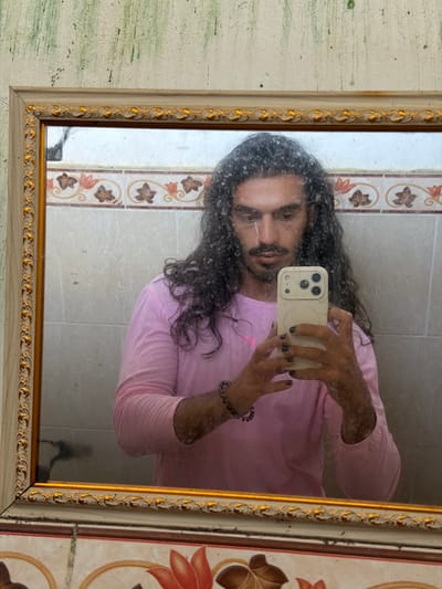 A person with long, dark, curly hair and a mustache is taking a selfie in a mirror. They are wearing a pink long-sleeved shirt. Their nails are painted black. They are holding a light-colored smartphone with a cream-colored case. The mirror has a decorative gold and cream frame and is positioned above a tiled surface with a floral pattern. The wall above the mirror appears to be textured and has some green marks. The scene is indoors, likely a bathroom.
