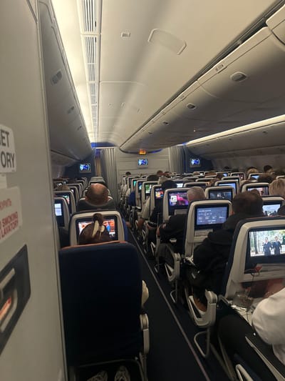 The image captures the interior of an airplane cabin, rotated 90 degrees clockwise. When viewed correctly, it shows a long airplane cabin with passengers seated, likely during a flight.
On the left side of the corrected image, the pristine white ceiling and overhead compartments of the aircraft are visible. The overhead bins are closed, featuring a sleek, modern design with subtle handles and ventilation grates along their lower edge. Recessed lighting strips run along the ceiling and beneath the bins, casting a soft, ambient glow.
On the right side, multiple rows of airplane seats stretch into the distance. Most passengers are seen from behind, settled into their seats. The seats are upholstered in a dark blue or black fabric. Each seatback is equipped with an individual in-flight entertainment (IFE) screen. Many passengers are actively engaged with these screens, which display various content, including what appears to be movies, TV shows, or flight information interfaces. Some screens show people in a wooded scene, while others display menu options. The screens have a light-colored bezel.
In the mid-ground, a passenger with a bald head is visible, looking at their screen, which appears to be showing a brightly colored animation or cartoon. Further back, another passenger is wearing a dark hoodie, and brown shoes are visible beneath their seat. The general activity is passive entertainment, typical of a long-haul flight.
The aisle runs down the center of the visible cabin section, separating the rows of seats from the overhead bins and providing a pathway through the aircraft. The floor is covered with a dark carpet.
The overall lighting in the cabin is dim, suggesting that it might be a nighttime flight or that the cabin lights have been lowered for passenger comfort. Small blue accent lights are visible further down the aisle in the distance, possibly marking exits or other facilities.
At the bottom of the original image (which would be in the foreground, near the floor, if corrected), partially visible text is present on what appears to be a sign or a safety card. The legible text includes "...ET TORY" and, below it, "NO SMOKING LAVATORY". The visible part of the sign appears to be white with black lettering.