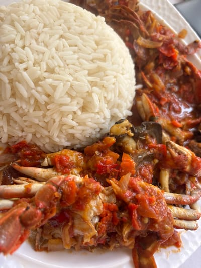 A close-up overhead shot captures a hearty meal served on a clean white plate. The plate is divided, with a generous, neatly mounded portion of fluffy white long-grain rice on the left side. Adjacent to the rice, filling the right side of the plate, is a dish of stewed crab in a rich, vibrant reddish-orange sauce.

The stew features multiple pieces of cooked crab, including visible claws and sections of the shell, thoroughly coated in the thick, savory sauce. Large, distinct chunks of red tomatoes and translucent slices of onions are clearly visible throughout the stew, suggesting a flavorful and aromatic preparation. The rice grains are separate and well-cooked, contrasting with the moist, saucy texture of the crab dish.

The scene focuses entirely on the prepared food, ready for consumption. The background is softly blurred, ensuring the meal is the central subject. There are no people visible, nor any discernible text. Given the provided context of Iwofe, Nigeria, this dish of stewed crab with rice is indicative of a local culinary offering, possibly a popular seafood meal from the region.