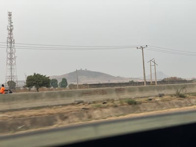 A hazy landscape features a cell tower, utility poles, and a distant hill. A concrete barrier runs horizontally across the foreground, separating the viewer from a field of dry vegetation. Several trees dot the landscape. A person in an orange shirt sits on the side of the road. The sky is overcast.