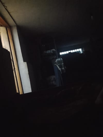 The image depicts a dimly lit interior, possibly a storage area or an unfinished room. A pale, rectangular pillar or wall feature dominates the left side of the frame, with some wooden elements visible beyond it, suggesting an opening or doorway. In the mid-ground, a figure is partially visible, wearing a horizontally striped shirt over a darker garment. The figure appears to be standing or leaning. Further into the background, a window or opening with horizontal bars is visible, letting in some light that illuminates a cluttered area. The overall atmosphere is dark and somewhat mysterious, with limited detail discernible due to the low light conditions. There is no visible text in the image. Given the location context of Kuk-Saray, Uzbekistan, this could be a room within a traditional dwelling, a historical building, or a storage space associated with local crafts or agriculture. The time of day cannot be determined, and no specific weather conditions are apparent from the interior scene.