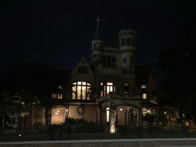 A nighttime photograph shows Mille Fleurs, one of the Magnificent Seven buildings located in Port of Spain, Trinidad and Tobago. The image is captured from across the street, with the wrought-iron fence and low brick wall in the foreground. The building itself is illuminated, revealing its unique architectural style. The lights from within the building cast warm glows through large windows, while the exterior is lit to highlight its towers, arches, and ornate details. Trees frame either side of the image, adding a sense of depth and enclosure to the scene. Above, the night sky is dark, with a few faint shapes suggesting distant aircraft.